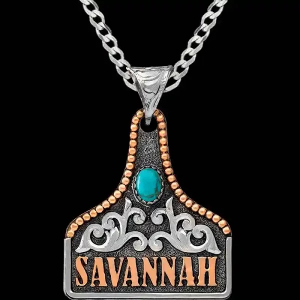 A matted silver cow tag necklace featuring copper lettering, silver scrollwork and turquoise stone